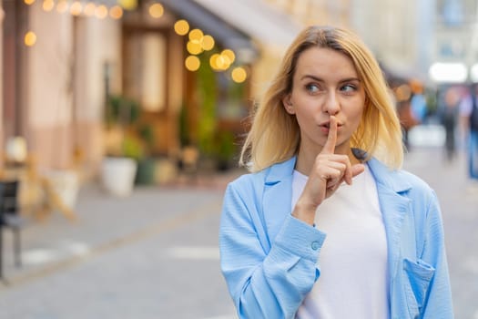 Shh be quiet please. Blonde young woman presses index finger to lips makes silence hush gesture sign do not tells gossip secret outdoors. Young lady female stand on urban city street. Town lifestyles