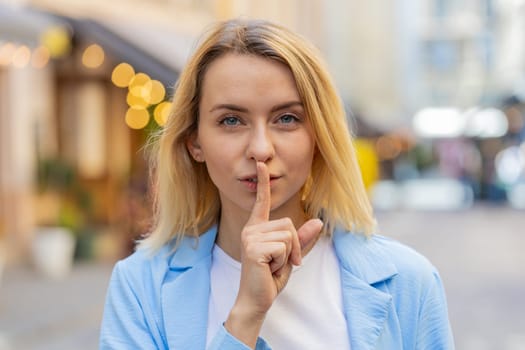 Shh be quiet please. Blonde young woman presses index finger to lips makes silence hush gesture sign do not tells gossip secret outdoors. Young lady female stand on urban city street. Town lifestyles