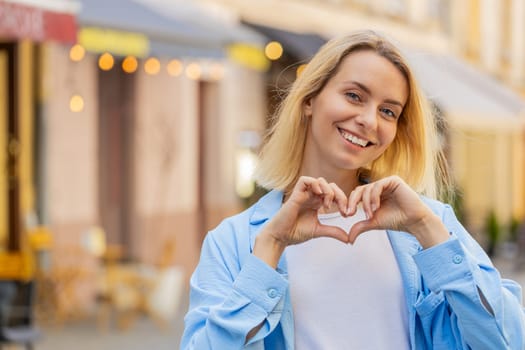 I love you. Caucasian woman makes symbol of love, showing heart sign to camera, express romantic feelings express sincere positive feelings. Charity, gratitude, donation. Outdoors in urban city street