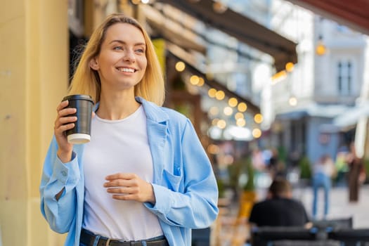 Happy Caucasian adult woman enjoying morning coffee hot drink and smiling. Relaxing, taking a break. Young lady standing on urban city center street, drinking coffee to go. Town lifestyles outside.