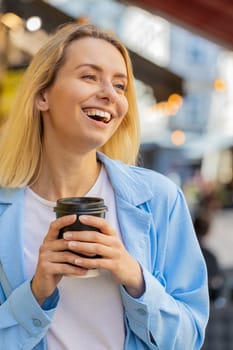 Happy Caucasian adult woman enjoying morning coffee hot drink and smiling. Relaxing, taking a break. Young lady standing on urban city center street, drinking coffee to go. Town lifestyles outside.