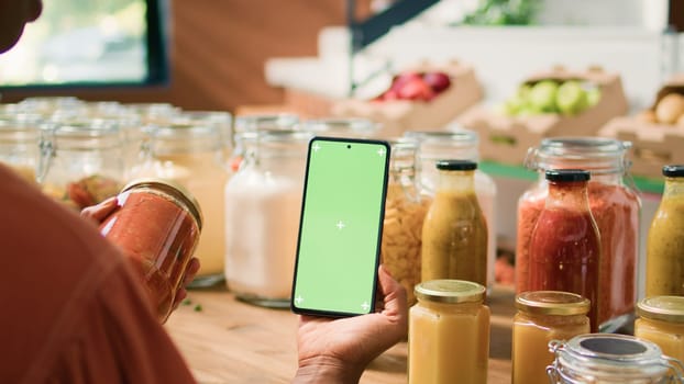 African american client uses greenscreen while she is looking at organic eco bulk products stored in reusable jars. Woman customer checks smartphone with isolated chromakey mockup display.