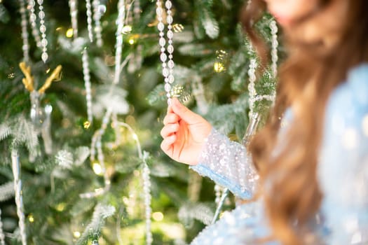 The girl decorates the Christmas tree, holding Christmas baubles in her hand. She wears a blue dress with a bow in her hair. Holiday concept