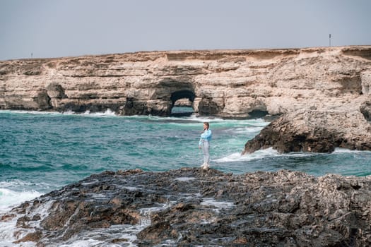 A woman in a blue jacket stands on a rock above a cliff above the sea and looks at the raging ocean. Girl traveler rests, thinks, dreams, enjoys nature. Peace and calm landscape, windy weather