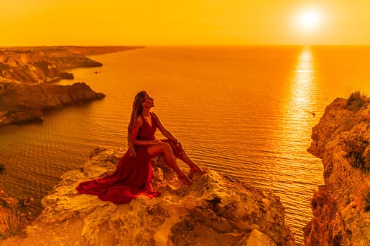 Woman sunset sea red dress, side view a happy beautiful sensual woman in a red long dress posing on a rock high above the sea on sunset