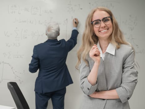 An elderly man writes on a white board and a young woman stands thoughtfully