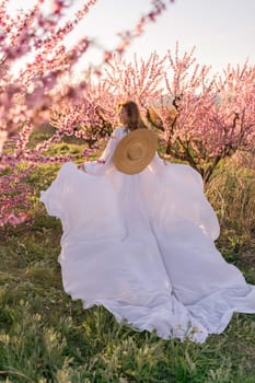 Woman blooming peach orchard. Against the backdrop of a picturesque peach orchard, a woman in a long white dress and hat enjoys a peaceful walk in the park, surrounded by the beauty of nature