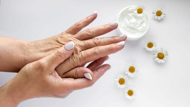 Close up of middle age woman hands applying cream with chamomile flowers and hand cream container on white background. Skincare and natural beauty concept. For healthcare, wellness, organic product.