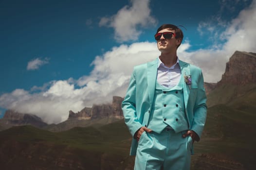 A groom in a suit stands against a background of mountains and clouds in summer. A man in a turquoise three-piece suit stands confidently looking to the side.