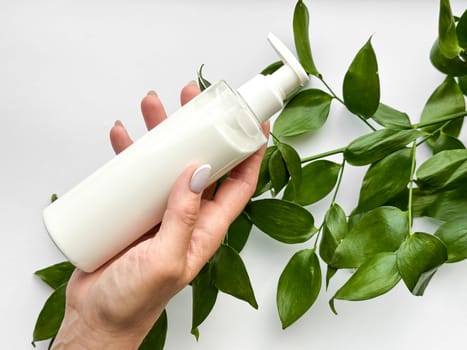 Close up of middle age woman hand holding white lotion bottle with green leaves on white background. Skincare and natural beauty concept. Healthcare, wellness, and organic product poster. High quality