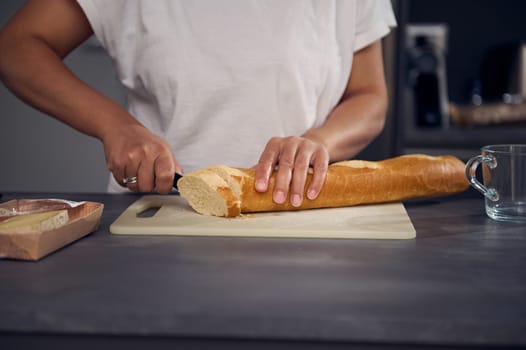 Close-up woman's hands using kitchen knife, cutting slicing loaf of bread a French baguette on cutting board, preparing toasts of sandwiches for breakfast at minimalist home kitchen interior