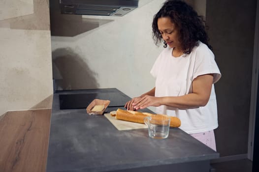 Side portrait of a woman using kitchen knife, cutting a loaf of whole grain French baguette bread on the cutting board, preparing delicious sandwiches with cheese for breakfast in the home kitchen.