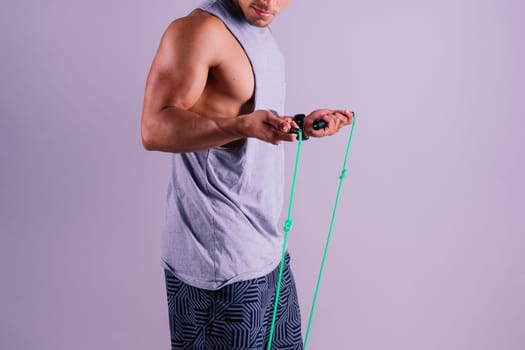 A man with a grey tank top is holding a green jump rope with his elbow bent and waist twisted, showing off his toned chest and wrist strength