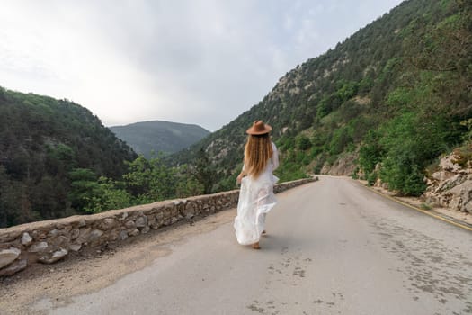 A woman in a white dress is walking down a road with a hat on. The road is surrounded by mountains and the sky is cloudy. The woman is enjoying the scenery and the peacefulness of the area