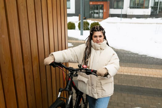 A woman with dreadlocks is standing next to a bicycle. She appears to be looking at something off-camera, her hands casually resting on the handlebar. The background shows a street setting with buildings.