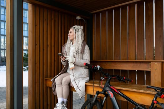 A woman is sitting on a wooden bench placed next to a parked bicycle. The woman is relaxed, looking around, possibly taking a break from cycling.