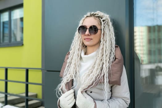 A woman with white dreadlocks leaning against a wall in an urban setting. She is dressed casually, with a contemplative expression on her face.