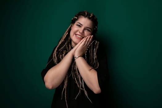 A woman with dreadlocks is standing upright in front of a bright green wall. She appears confident and is looking straight ahead. The contrast between her dark hair and the vivid green background is striking.