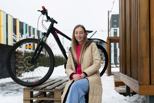 A woman is seated on a wooden bench, positioned next to a bicycle. The woman appears relaxed and is potentially taking a break. The bike rests against the bench, suggesting it is her mode of transportation.