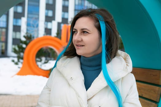 A woman with vibrant blue hair sits on a wooden bench in a public park. She is dressed in casual clothing, with headphones in her ears, looking off into the distance.