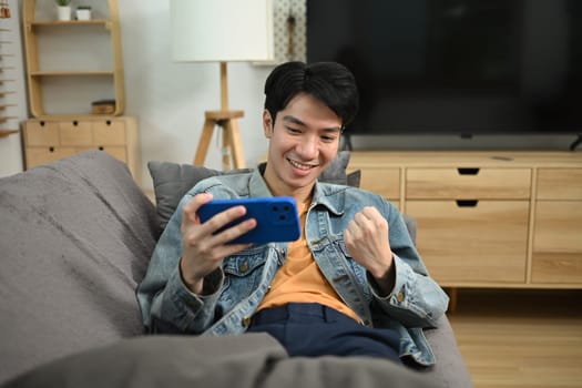 Happy asian man sitting on couch and celebrating victory in mobile game with raised fist.