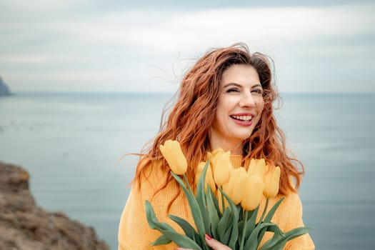 Portrait of a happy woman with hair flying in the wind against the backdrop of mountains and sea. Holding a bouquet of yellow tulips in her hands, wearing a yellow sweater.