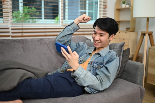 Young man relaxing on couch at home and looking at cellphone celebrating online prize win.