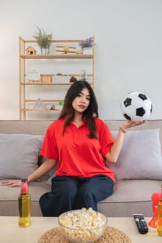 Asian woman holding soccer ball at home with snacks and drinks. Concept of sports, leisure, and relaxation.