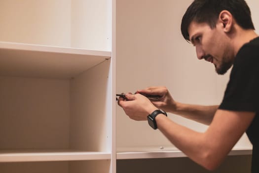 Young handsome caucasian brunette man is screwing a screw with a screwdriver on a metal fitting inside a closet while standing in the bedroom, close-up side view with selective focus. Furniture assembly concept.