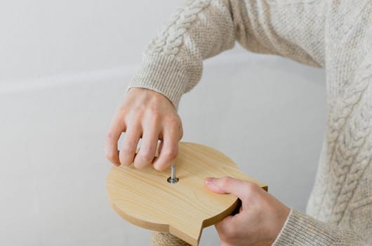Hands of a young caucasian man in a knitted sweater manually twists a screw with a curly screwdriver into a board while assembling a cat scratching post against a white wall, top view close-up. Concept assembly of a scratching post, cat furniture.