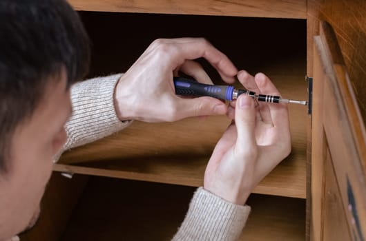 One young Caucasian man manually tightens a bolt into the door of an old wooden cabinet with a screwdriver, side view close-up with selective focus.