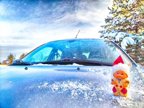 Cute little funny dwarf in the snow on hood of the car in winter landscape. The concept of winter travel. Background for Christmas and New Year