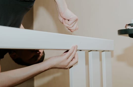 One young Caucasian unrecognizable man bending down puts a screw through a hole in the white side of a bunk bed, in the evening in the room, side view close-up with selective focus.