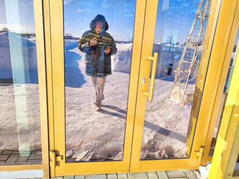 Woman Capturing Her Reflection in Snowy Doorway. A woman takes a reflective selfie amidst winter scenery