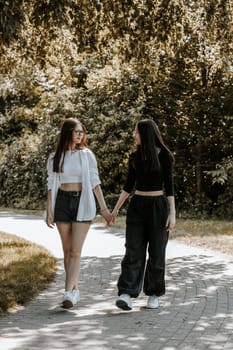 Portrait of two young beautiful Caucasian girls holding hands and looking at each other in the eyes stroll along the path in a city park on a summer day, close-up view from the side.