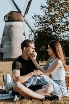 One beautiful Caucasian brunette girl brings an apple to her boyfriend s mouth wanting to treat him, sitting romantically sideways on a blanket with a basket in the park on a picnic against the backdrop of an old mill, close-up side view.