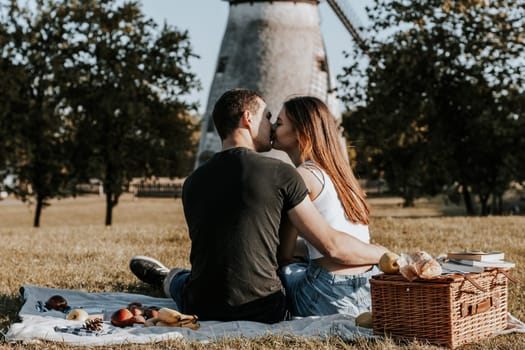 One beautiful Caucasian guy hugging a girl and gently kissing her on the lips on a sunny spring day, sitting with their backs on a blanket with a basket, books and fruits in the park on a picnic against the backdrop of an old mill, close-up view from below.