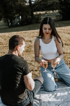 One beautiful Caucasian brunette girl with long flowing hair with a smile holds glasses of champagne in her hands and serves them to a guy sitting with his back turned in the park on a picnic, close-up side view.