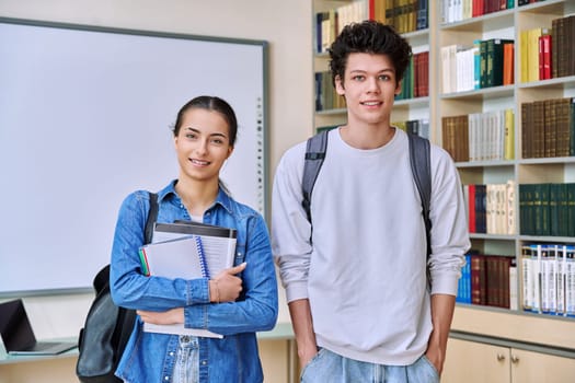 Portrait of two teenage college students classmates, guy and girl, inside library of educational building. Smiling teenage friends looking at camera together. Education, adolescence, youth, lifestyle