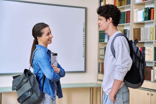 Portrait of two teenage college students classmates, guy and girl, inside library of educational building. Smiling teenage friends talking together. Education, adolescence, youth, lifestyle concept