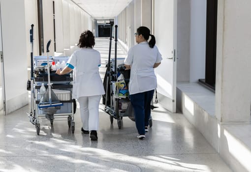 Two female cleaners with cleaning carts walk along a long white corridor. Cleaning concept in office and hospital.