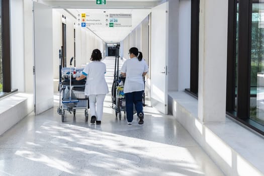 Two female cleaners with cleaning carts walk along a long white corridor. Cleaning concept in office and hospital.