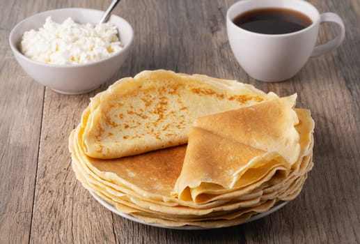 A stack of thin pancakes, a plate with cottage cheese and coffee on a wooden table.