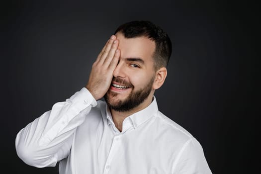 portrait of handsome bearded man closes his eyes with hand and spying on black background