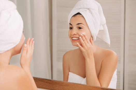 Portrait of beautiful woman caring of her skin. girl looking at mirror and applying cream on her face in front of the mirror in bathroom