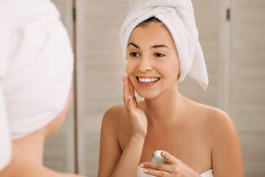 Portrait of beautiful woman caring of her skin. girl looking at mirror and applying cream on her face in front of the mirror in bathroom