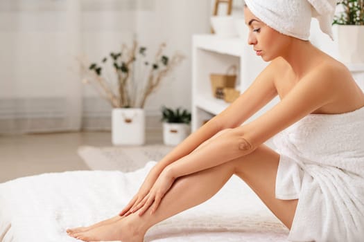 Young beautiful woman in towel applying cream on her legs after shower at home