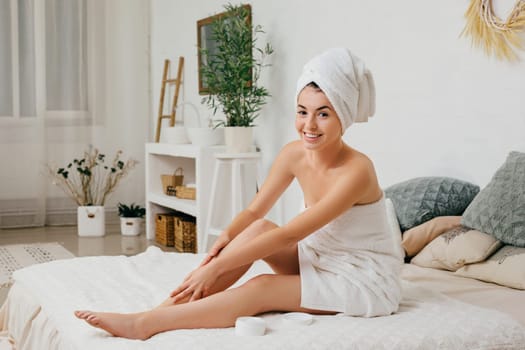 Young beautiful woman in towel applying cream on her legs after shower at home