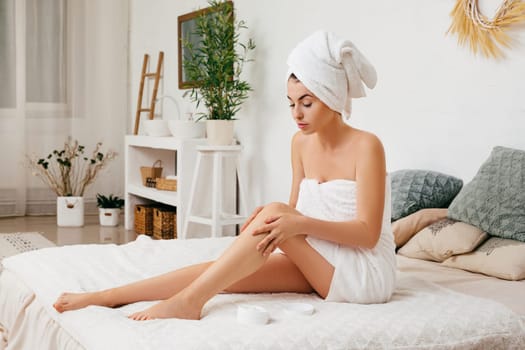 Young beautiful woman in towel applying cream on her legs after shower at home