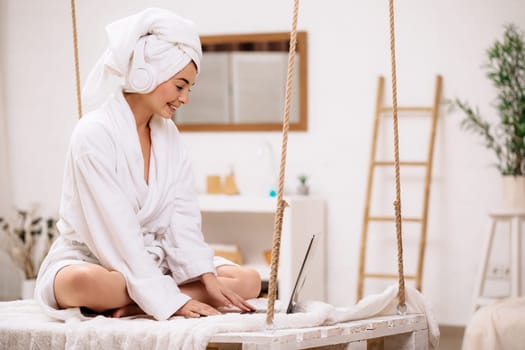 woman in bathrobe and towel after shower uses laptop in bedroom